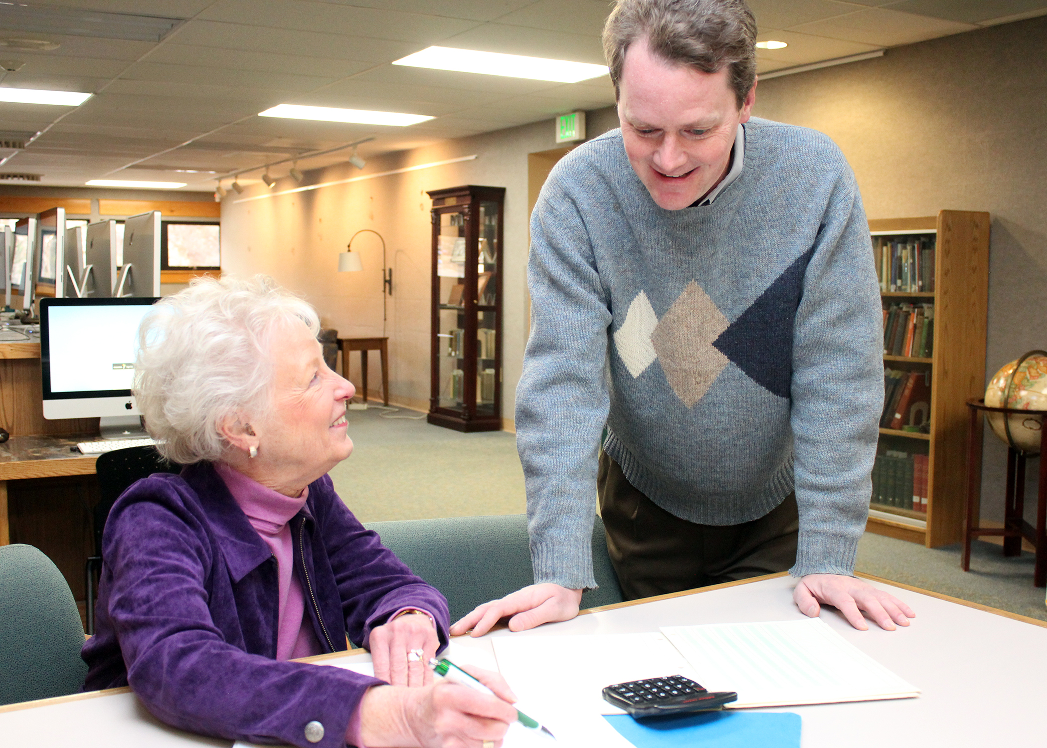 man and woman in library looking at ledger and calculator on desk